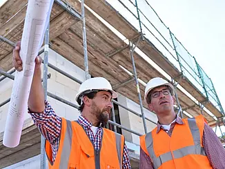 Zwei Bauleiter auf einer Baustelle mit Bauplan in der Hand bei der Abstimmung von Bauablauf, Terminen und Projektfortschritt.