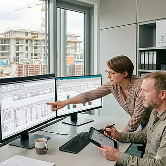 Zwei Experten im Büro analysieren einen digitalen Soll-Ist-Vergleich am Bildschirm mit Blick auf eine Baustelle im Hintergrund.
